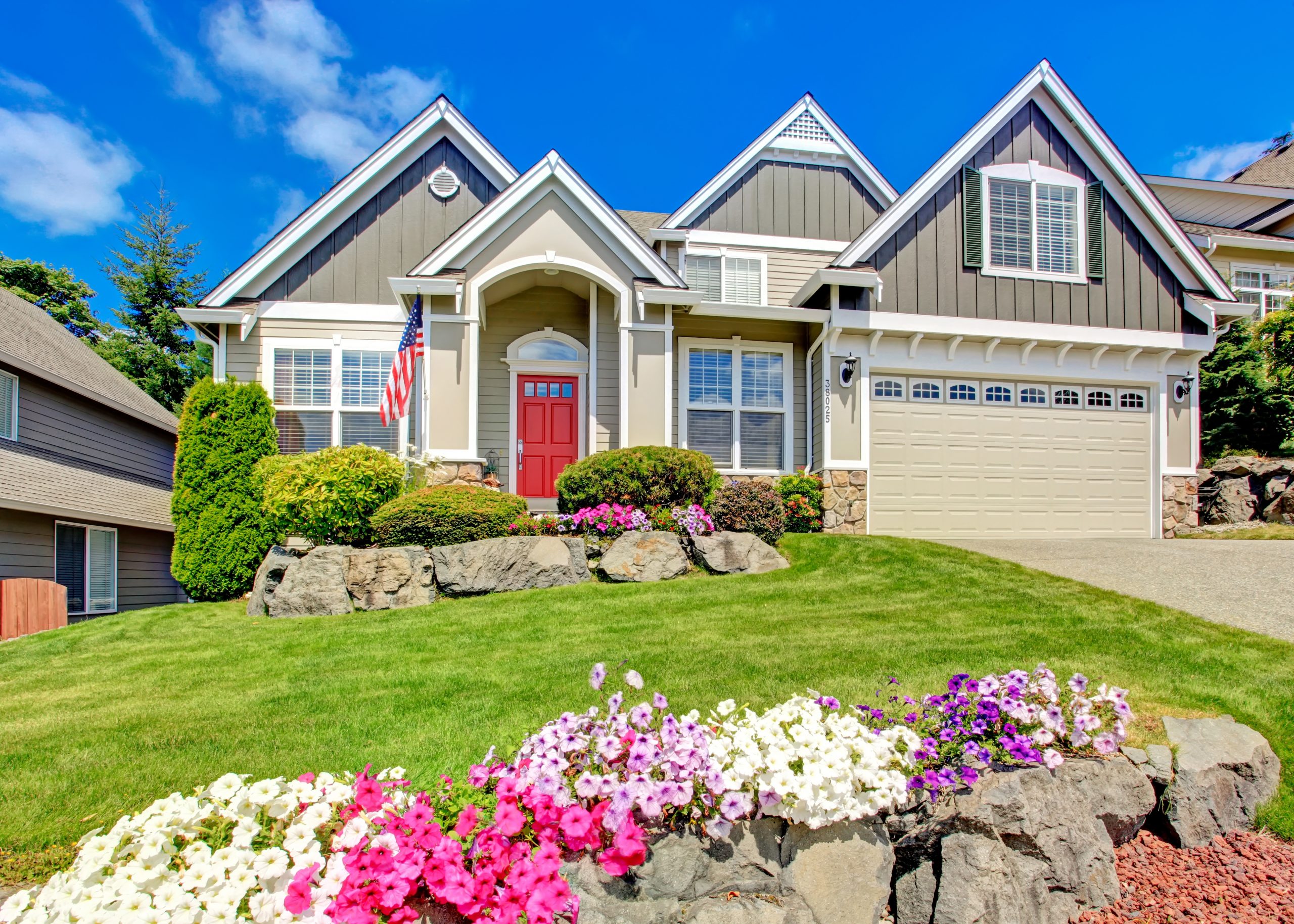 Grey house exterior with entrance porch and red door. Beautiful front yard landscape with vivid flower and stones