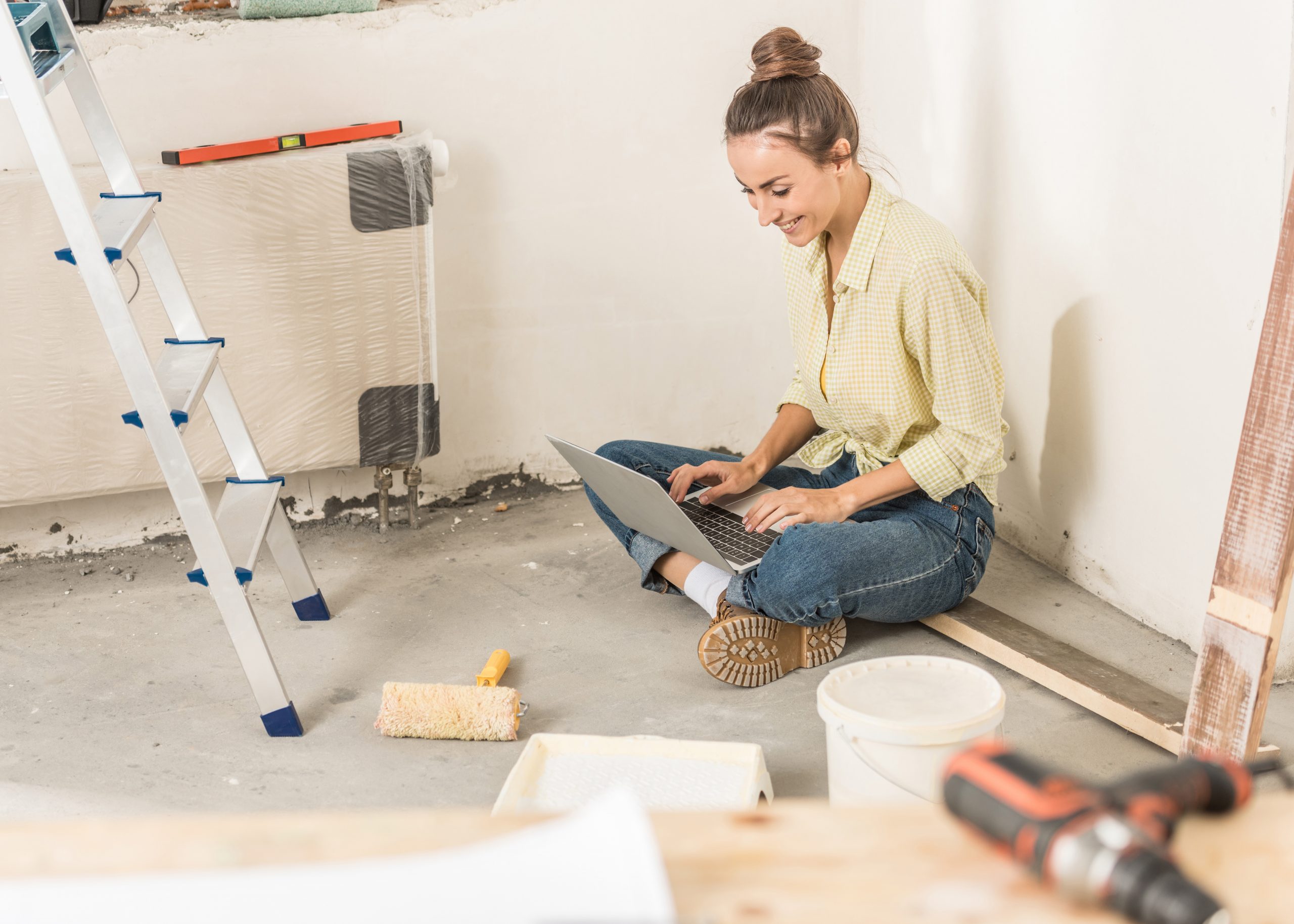 Young Caucasian woman sitting on the floor of a room being remodeled, using a laptop.