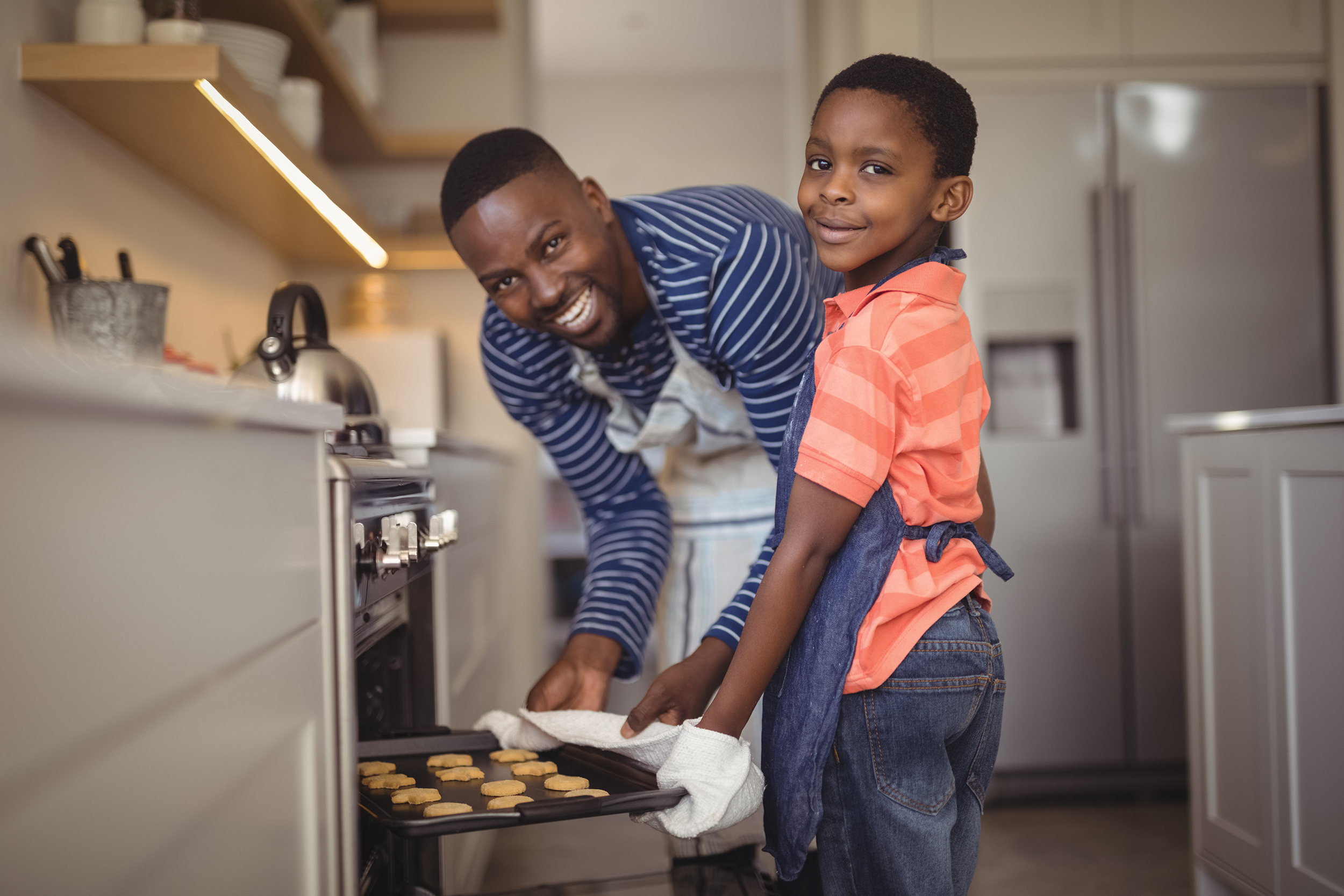 Father taking tray of fresh cookies out of oven with son in kitchen African American father and son in the kitchen baking cookies.